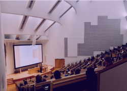 Auditorio, jonka katsomossa istuu ihmisiä. Puhuja seisoo lavalla ja lavan valkokankaalle on heijastettu esitys.